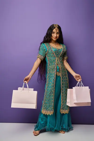 Tiro vertical de mulher indiana atraente em sari azul posando com sacos de compras em pano de fundo roxo — Fotografia de Stock