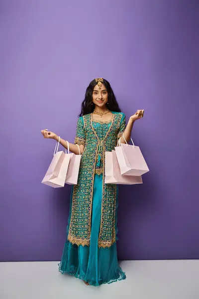 Alegre hermosa mujer india en traje nacional posando con bolsas de compras sobre fondo púrpura - foto de stock