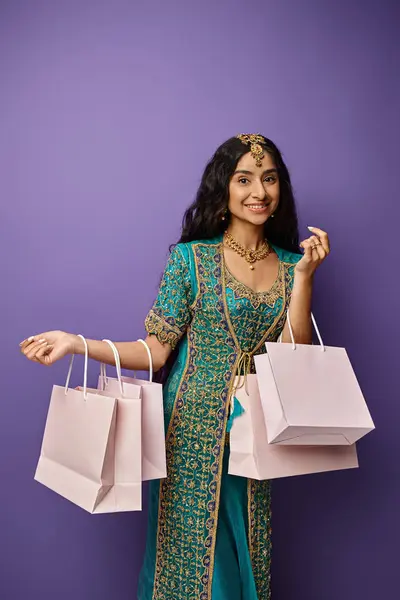 Jolly indian woman in traditional clothes with accessories holding shopping bags on purple backdrop — Stock Photo