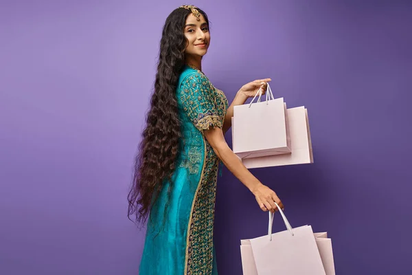 Jolie femme indienne joyeuse aux cheveux longs en sari bleu posant avec des sacs à provisions sur fond violet — Photo de stock