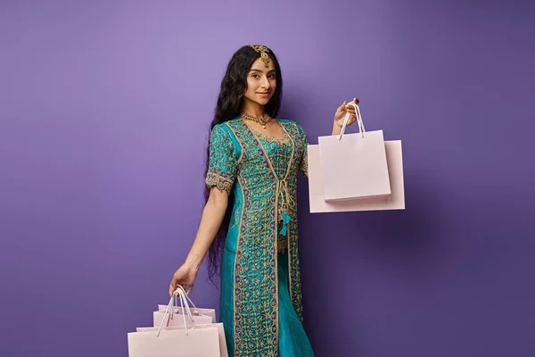 Schöne Indianerin in blauem Sari mit Accessoires, die Einkaufstaschen vor lila Hintergrund halten — Stockfoto