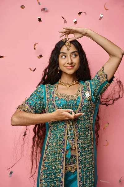 Jolly attractive indian woman gesturing and looking away under confetti rain on pink backdrop — Stock Photo