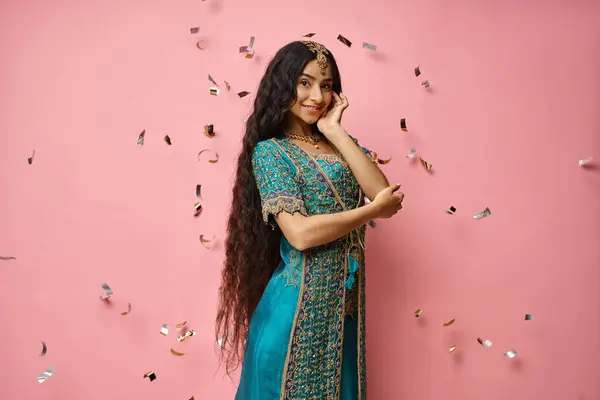 Allegra giovane donna indiana in sari blu guardando la macchina fotografica sotto la pioggia coriandoli su sfondo rosa — Foto stock