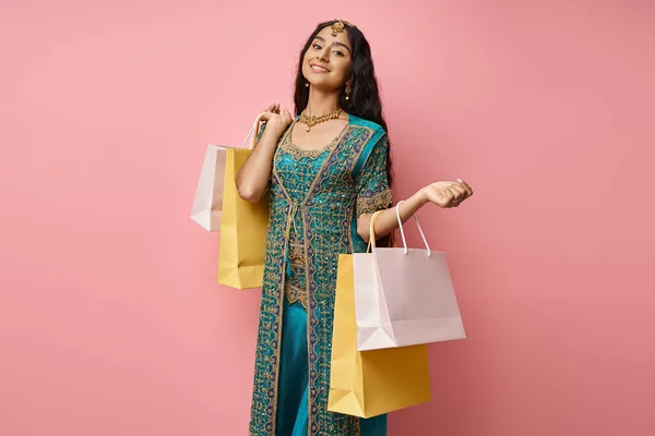 Cheerful young indian woman in national costume posing with shopping bags on pink background — Stock Photo