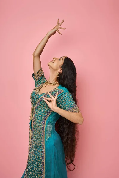 Tiro vertical de la atractiva joven india en sari azul gestos mientras baila sobre fondo rosa - foto de stock