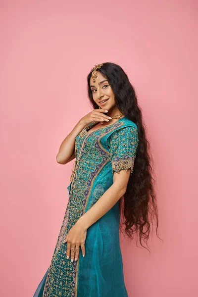 Tiro vertical de hermosa mujer india alegre en sari azul con accesorios posando sobre fondo rosa - foto de stock