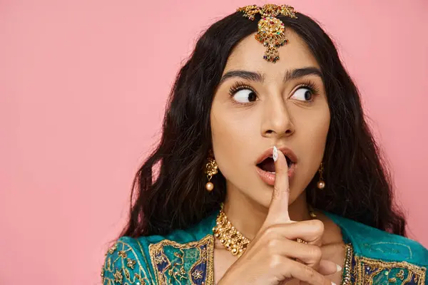 Shocked young indian woman in traditional costume posing with finger near mouth on pink backdrop — Stock Photo