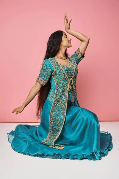 Young indian woman in national costume gesturing while dancing sitting on floor on pink backdrop — Stock Photo