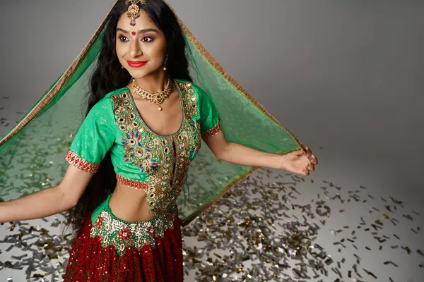 Joyeuse jeune femme indienne en costume national avec point bindi posant sur confettis et détournant les yeux — Photo de stock