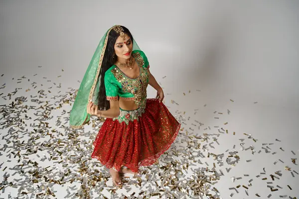 Joven mujer bastante india en ropa tradicional posando en confeti en el suelo y mirando hacia otro lado - foto de stock