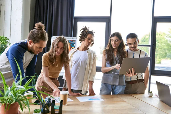 Creative multiethnic team discussing startup near laptop and graphs in modern office, brainstorming — Stock Photo