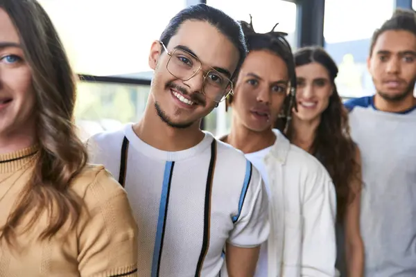 Headshot of glad team lead in eyeglasses looking at camera with multicultural colleagues in office — Stock Photo