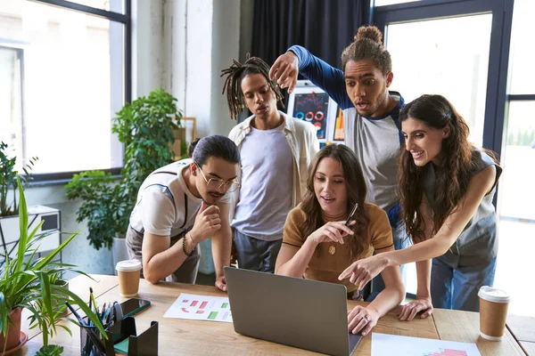 Équipe multiculturelle émerveillée et excitée pointant vers ordinateur portable près des graphiques sur le bureau, remue-méninges — Photo de stock