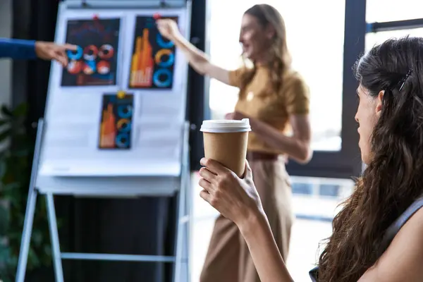 Businesswoman with coffee to go looking at colleagues talking near flip chart with graphs in office — Stock Photo