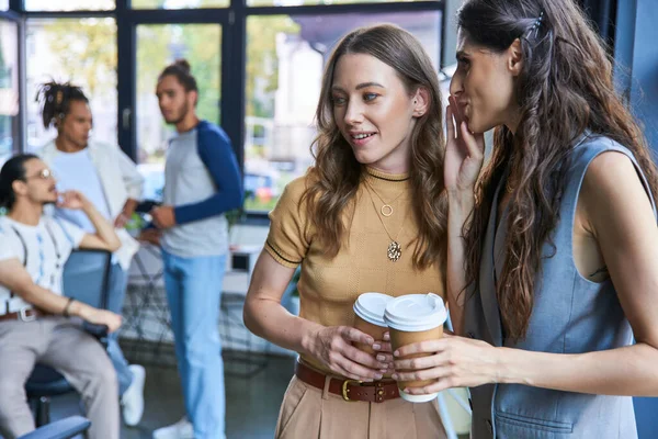 Empresária dizendo segredo para colega feminino durante coffee break perto de homens multiétnicos no escritório — Fotografia de Stock