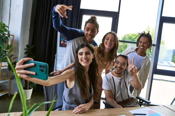 Mulher alegre com smartphone tomando selfie com equipe multiétnica gesticulando no escritório moderno — Fotografia de Stock