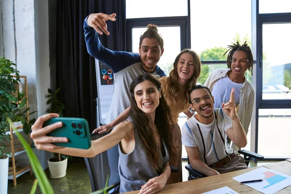 Mujer sonriente tomando selfie en el teléfono inteligente con colegas multiculturales felices en la oficina moderna - foto de stock