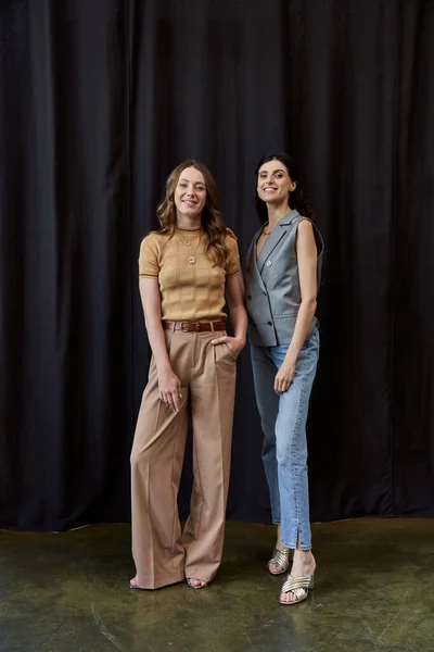 Full length of two joyful businesswomen in stylish casual attire standing near black drape in office — Stock Photo