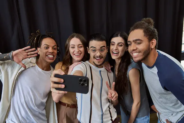 Joyful man in eyeglasses showing victory sign and taking selfie on smartphone with multiethnic team — Stock Photo