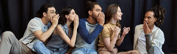 Interracial team sitting and telling secret to amazed colleague on black backdrop in office, banner — Stock Photo