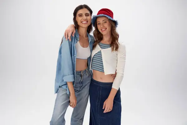 Femme joyeuse en panama chapeau embrassant petite amie en denim vêtements sur fond gris, mode de rue — Photo de stock