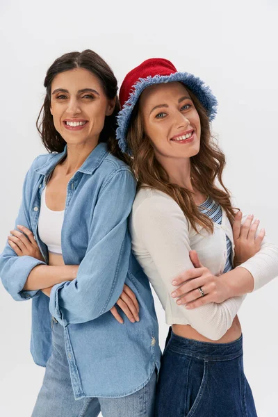 Cheerful women in jeans wear and panama hat posing back to back with folded arms on grey backdrop — Stock Photo