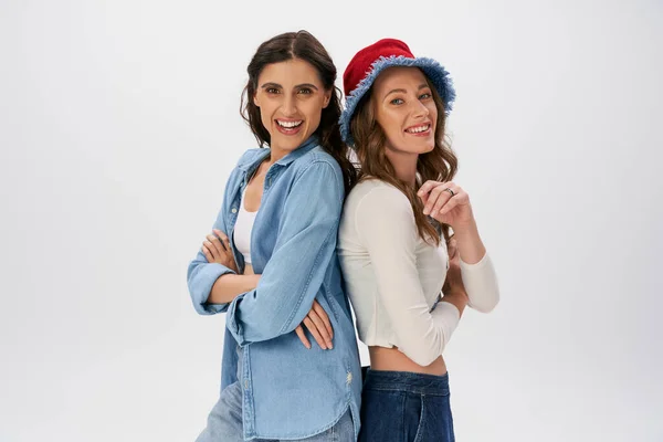 Happy women in jeans wear and panama hat posing back to back with folded arms on grey, fashion — Stock Photo