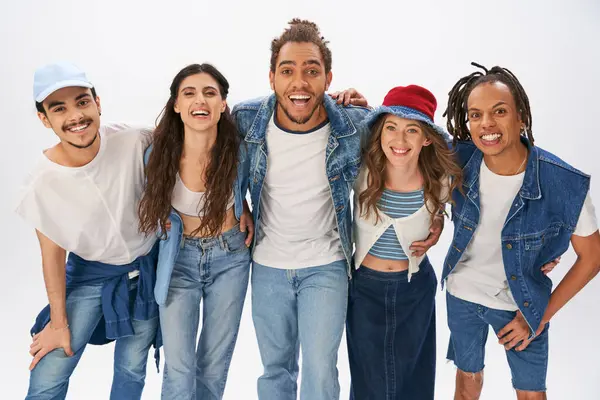 Carefree multicultural friends in trendy street wear embracing and smiling at camera on grey — Stock Photo