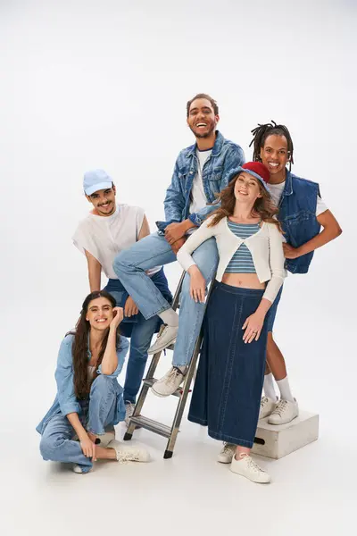 Joyful multicultural friends in trendy clothes posing with ladder and looking at camera on grey — Stock Photo