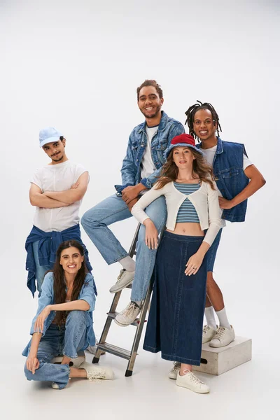 Heureux amis multiethniques dans les vêtements de rue à la mode posant avec échelle et souriant à la caméra sur gris — Photo de stock
