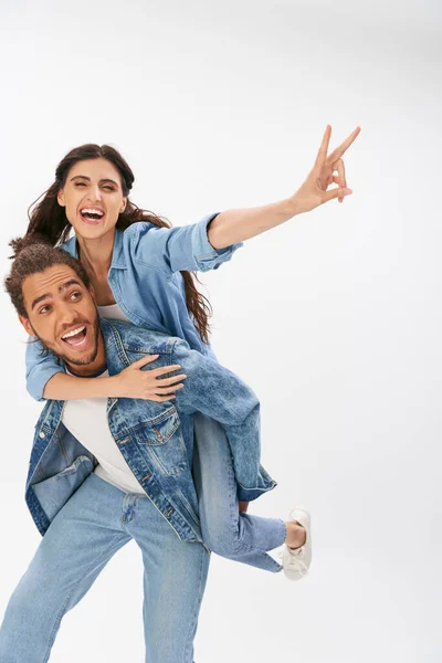 Cheerful african american man piggybacking girlfriend in denim clothes showing victory sign on grey — Stock Photo