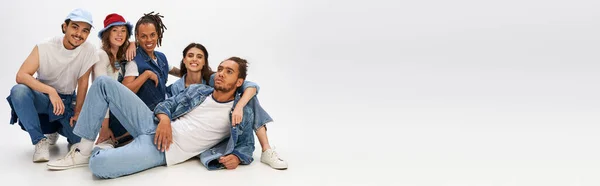 Trendy african man in denim clothes lying down near happy multiethnic friends posing on grey, banner — Stock Photo