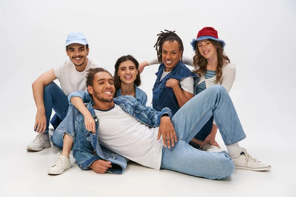 Fröhlicher afrikanischer Mann in Jeans in der Nähe von interrassischen Freunden liegend und in die Kamera blickend auf grau — Stockfoto