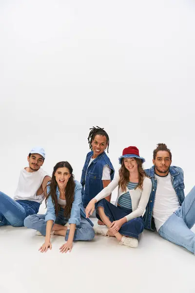 Amigos multiétnicos felices en ropa de moda y de mezclilla sentado y mirando a la cámara en el fondo gris — Stock Photo