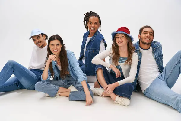 Stylish multiethnic friends in casual street wear sitting and smiling at camera on grey backdrop — Stock Photo