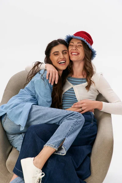 Cheerful and stylish girlfriends in denim outfits embracing in armchair on grey backdrop, fashion — Stock Photo