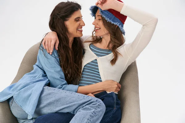 Fröhliche Freundinnen in trendiger Freizeitkleidung, die sich im Sessel vor grauem Hintergrund anschauen — Stockfoto