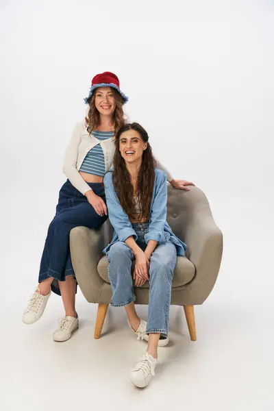 Cheerful woman in denim clothes sitting in armchair near stylish girlfriend on grey backdrop — Stock Photo