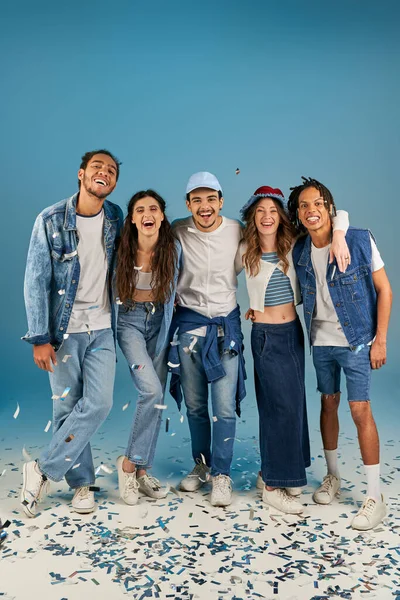Amis multiethniques à la mode en denim et mode porter rire près de confettis festifs sur bleu — Photo de stock
