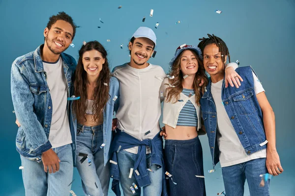 Amigos multiétnicos felizes em roupas casuais na moda sorrindo para a câmera sob chuva de confetes em azul — Fotografia de Stock
