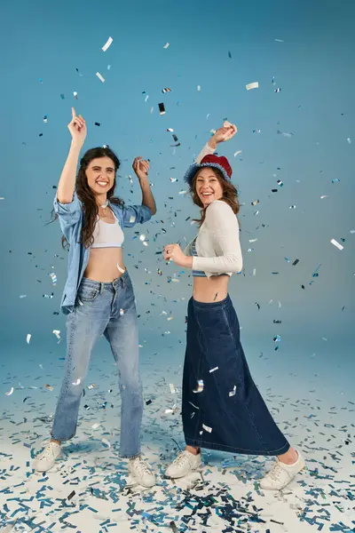 Full length of cheerful and trendy girlfriends under sparkling confetti rain on blue backdrop — Stock Photo