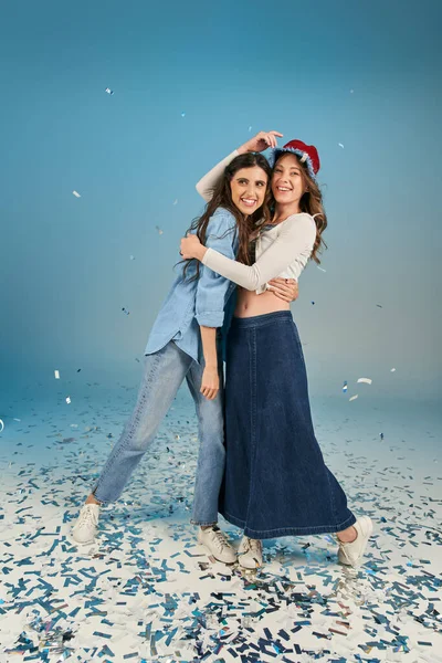 Полная длина радостных и стильных подруг, обнимающих на голубом фоне праздничные конфетти — стоковое фото