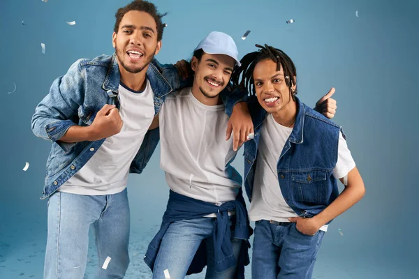 Unbekümmerte gemischtrassige Freunde in trendiger Jeanskleidung blicken unter glänzendem Konfetti auf blau in die Kamera — Stockfoto