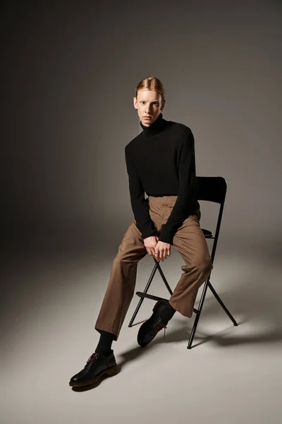 Atractiva persona no binaria en cuello alto negro y pantalones marrones sentados en silla negra, moda - foto de stock