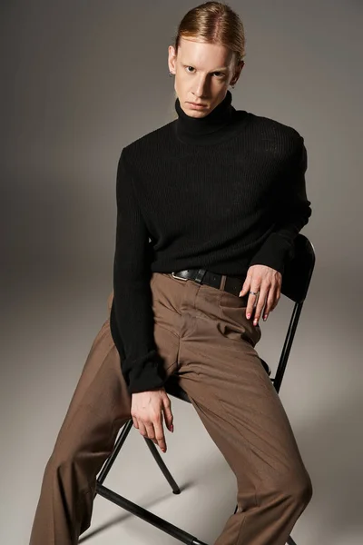 Good looking young non binary person with ponytail sitting on black chair and looking at camera — Stock Photo