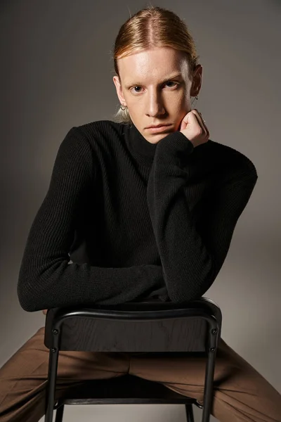 Приваблива молода небінарна людина, що сидить на чорному стільці з рукою під підборіддям, концепція моди — стокове фото