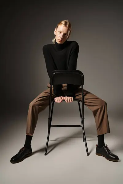 Atractivo no binario persona en b falta cuello alto y pantalones marrones sentado en silla negra, moda - foto de stock