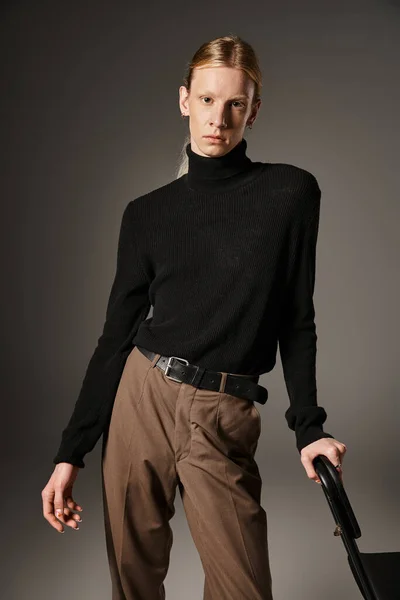 Vertical shot of young non binary person with ponytail posing with hand on black chair, fashion — Stock Photo