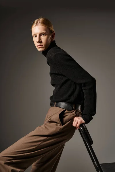Attractive young non binary person in black turtleneck leaning on black chair, looking at camera — Stock Photo