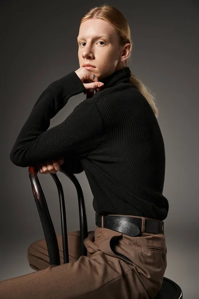 Vertical shot of young non binary model posing on black chair with hand under chin looking at camera — Stock Photo